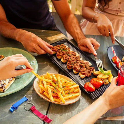 A group of people sharing a meal at an outdoor dining table, with a black rectangular platter holding stuffed bread slices topped with meat sauce, a plate of french fries, tomatoes, and other dishes. Two colorful keychains are visible on the table.
