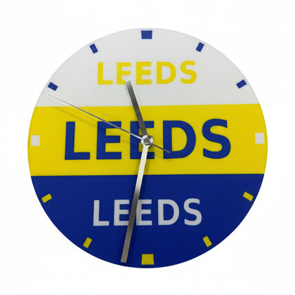 A circular wall clock featuring the Leeds club colors, with a white top half, yellow middle band, and blue bottom half, all printed with the word 'LEEDS'. The clock has silver hands and small blue and white hour markers.
