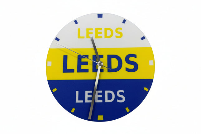 A circular wall clock featuring the Leeds club colors, with a white top half, yellow middle band, and blue bottom half, all printed with the word 'LEEDS'. The clock has silver hands and small blue and white hour markers.