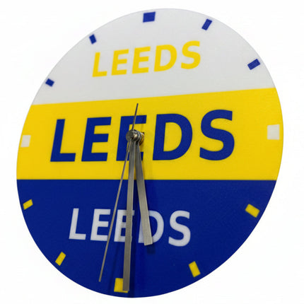 A circular wall clock featuring the Leeds United FC logo colors and text, with 'LEEDS' printed across white, yellow, and blue sections. The clock has silver hands and no visible brand logo beyond the team text.