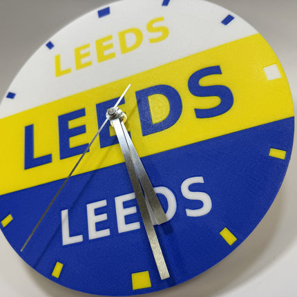 A circular wall clock featuring the Leeds United football club logo colors and text, with silver hands on a plain light background.