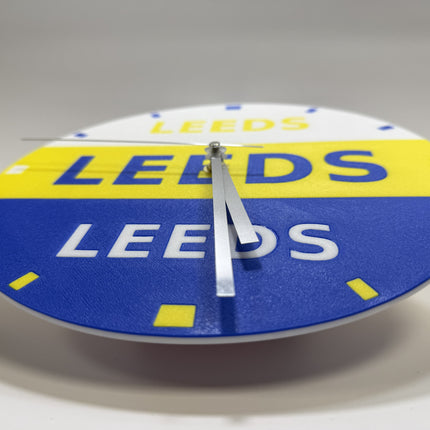 A round wall clock featuring the Leeds United FC colors, with a white top section, a yellow band, and a blue lower section, all printed with the word 'LEEDS'. The clock has silver hands and no visible brand logo.