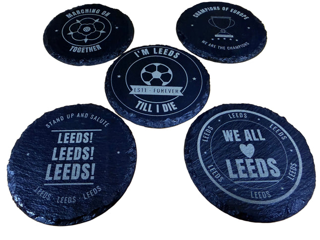 Five round black slate coasters with white printed football-themed designs and text, arranged on a light wood surface.