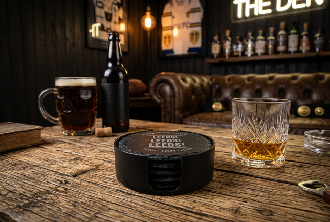A black circular coaster with 'LEEDS LEAGUE 2025' printed on it, styled on a rustic wooden bar table alongside a bottle of beer, a mug of dark beer, a cut glass of whiskey, and a bottle opener, with a bar interior in the background.